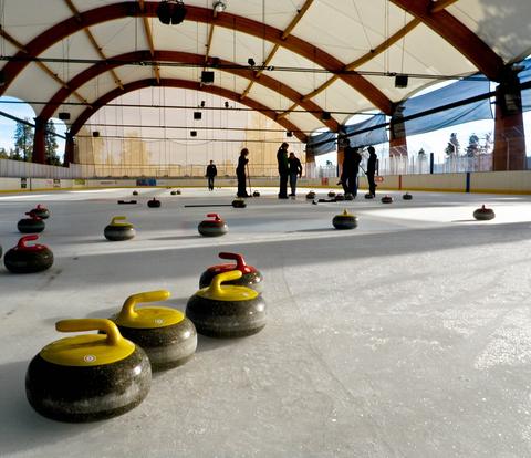 Yellow and red curling stones resting on an indoor ice sheet with players in the background, curling