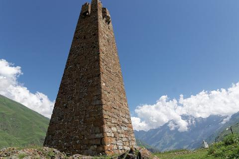 Tall weathered stone defensive tower rising against a blue sky and mountains, Turret Siege